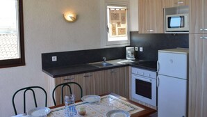 A compact kitchen with a sink, stove, microwave, and a small refrigerator. There is a window with a decorative view, a wall-mounted light, and a table with chairs.