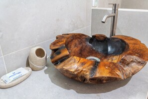Ein einzigartiges, rundes Holzbecken mit einem modernen Wasserhahn, platziert auf einem gefliesten Boden neben einem Steinkübel und einem kleinen weißen Schild.