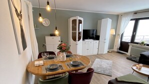 Un comedor con una mesa redonda, sillas y un reloj en la pared. Hay un aparador con puertas de cristal y un televisor al fondo.