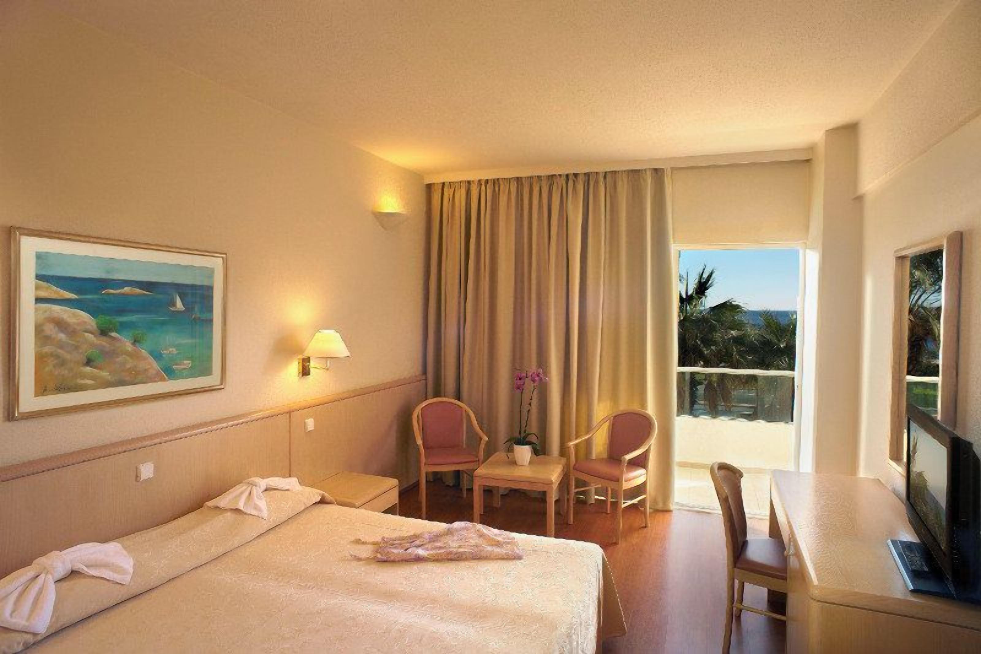 A hotel room with a bed, two chairs, a desk, a TV, and a painting.