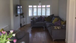 Una sala de estar con un sofá gris, una mesa de centro y un jarrón de tulipanes. Se ve una ventana con persianas y un candelabro.