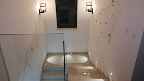 Una escalera con barandillas de cristal, una ventana con marco oscuro y luces fijadas a la pared.