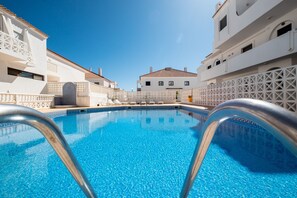 Une piscine à l’eau d’un bleu limpide, entourée de bâtiments blancs dotés de balcons, sous un ciel dégagé.
