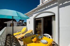 Un espace de restauration extérieur baigné de lumière, avec une table, des chaises, un parasol bleu et une porte vitrée donnant sur un espace intérieur.
