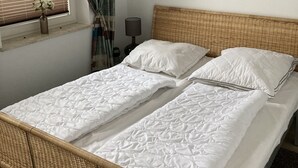 Un lit double avec une literie blanche, une tête de lit en osier, une fenêtre avec des stores, une lampe fixée au mur et un tableau floral encadré.