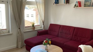 Un salon avec un canapé rouge, une table basse en bois sur laquelle se trouvent un livre et des fleurs, une fenêtre avec des rideaux et une bibliothèque remplie de livres et d’un téléphone.