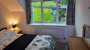 Un dormitorio con cama, mesita de noche, lámpara, ventana con cortinas y vista a un jardín.