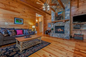 A cozy log cabin interior with a stone fireplace, wooden floors, a sofa with patterned cushions, and a ceiling fan. - Nature's Den | Pet-Friendly, Mountaintop Cabin in Columbus (Columbus)