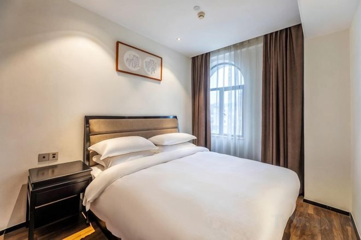A hotel room with a large bed, a bedside table, a wall-mounted light, and a window with curtains.