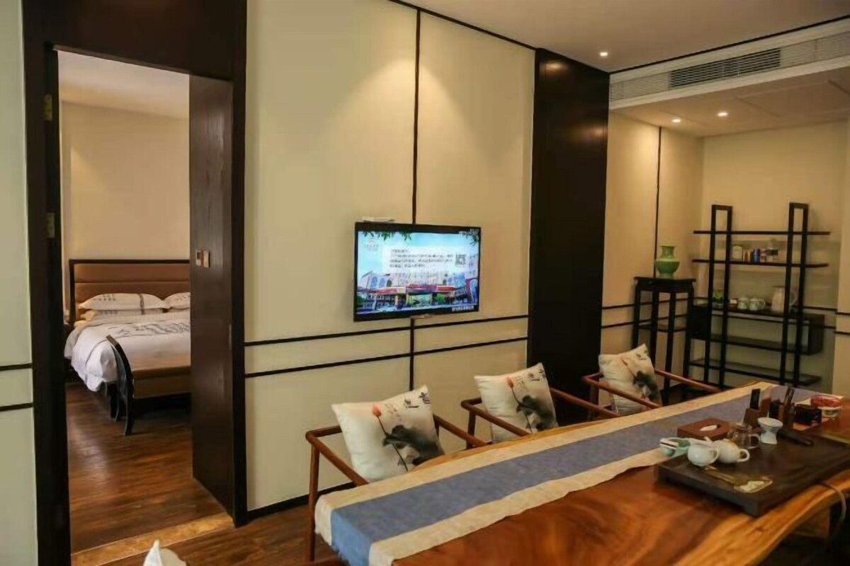 Una habitación de hotel con cama, televisión montada en la pared, una mesa de comedor de madera con sillas y estante con adornos.