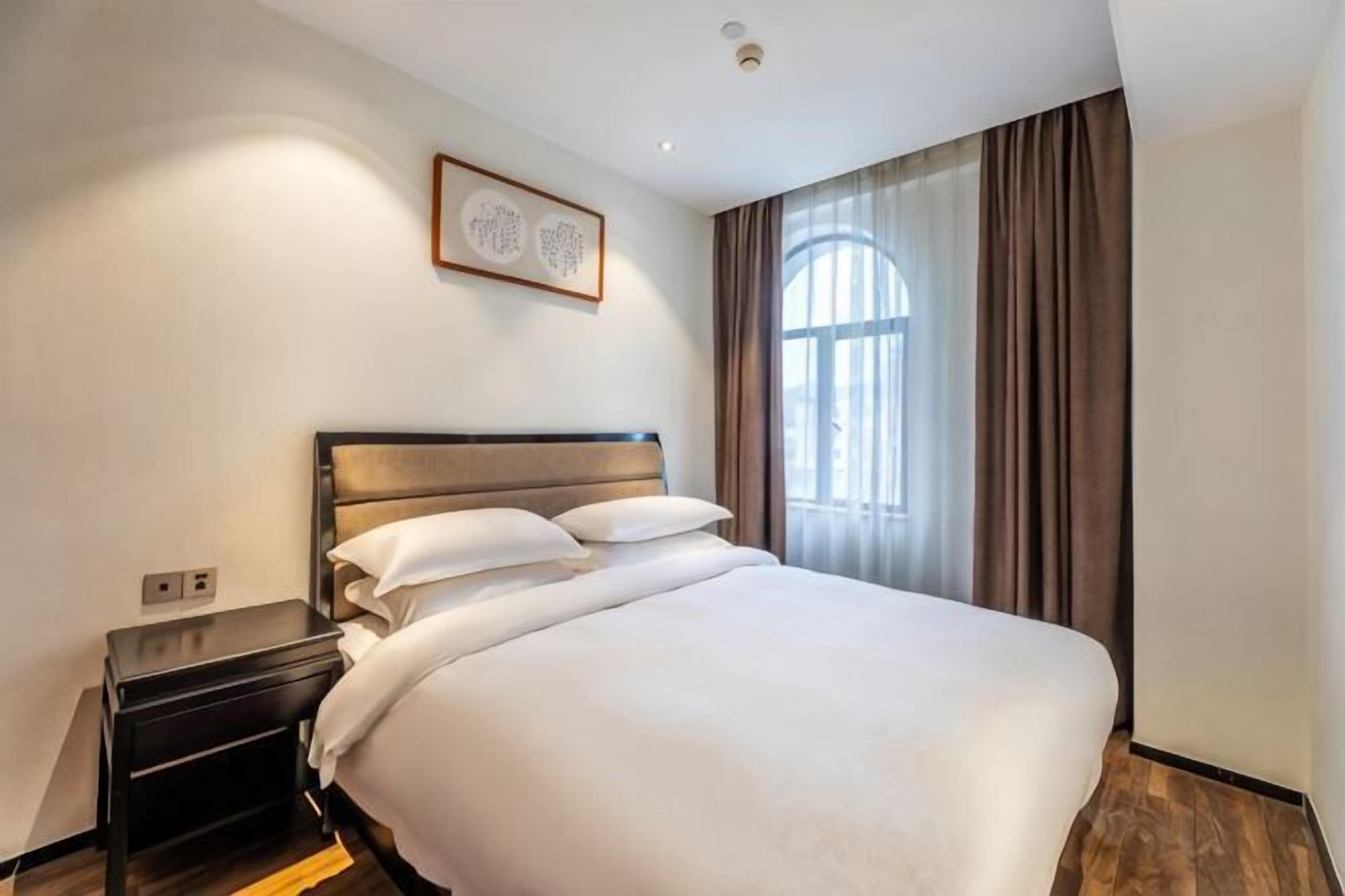 Ein Hotelzimmer mit einem großen Bett, einem Nachttisch, einem Wandbild und einem Fenster mit Vorhängen.