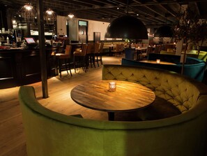 A modern bar area with a wooden bar counter, multiple bar stools, and a seating area with a round table and cushioned bench.