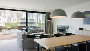 A modern living room with a dining area, large windows, and a view of a water body and buildings. - Luxury Villa With Jetty, at the Veerse Meer (Arnemuiden)
