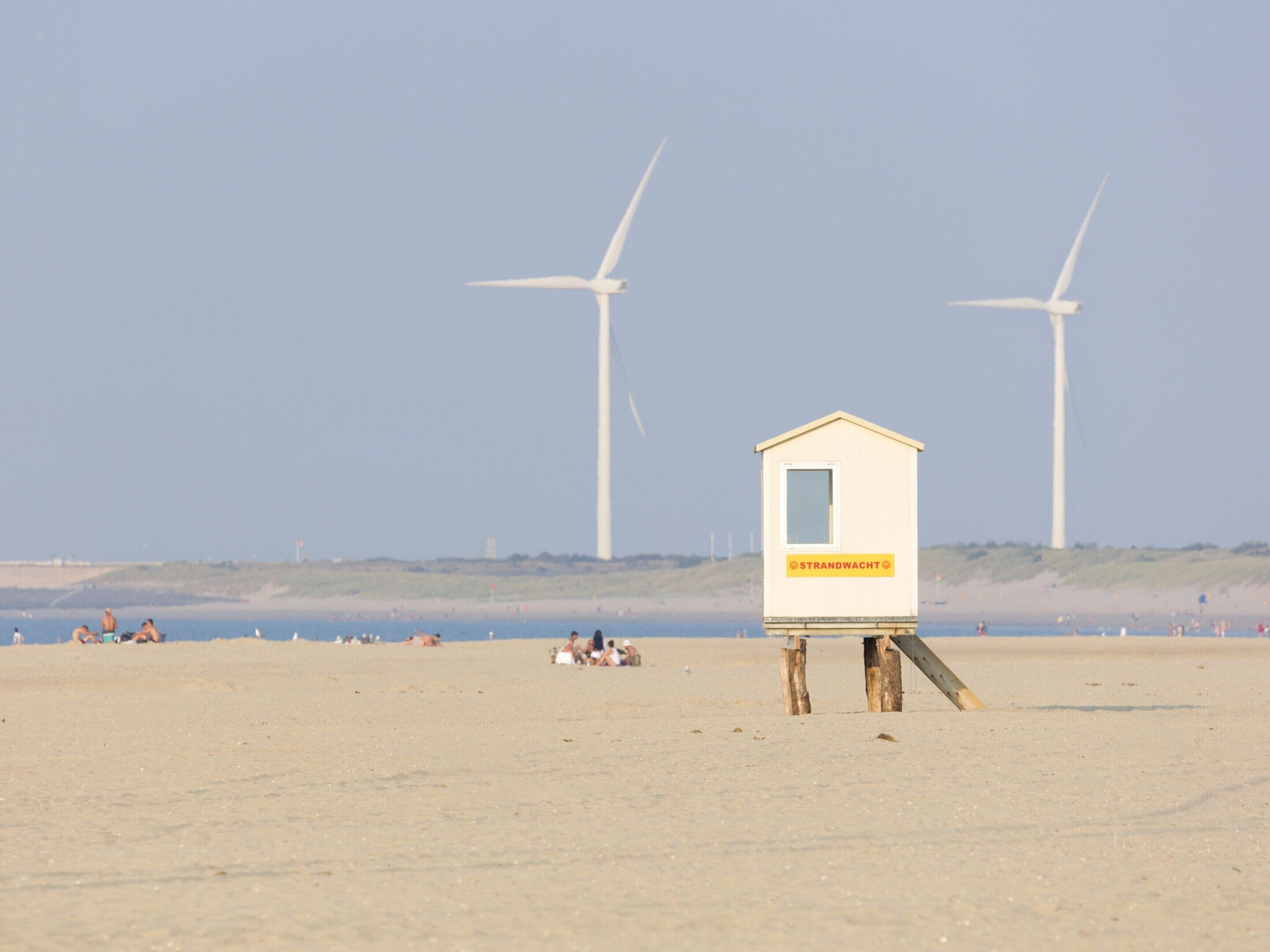 Ein Strand mit einem Rettungsschwimmturm, der 'STRANDWACHT' zeigt, und zwei Windkraftanlagen im Hintergrund.