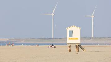 Ein Strand mit einem Rettungsschwimmturm, der 'STRANDWACHT' zeigt, und zwei Windkraftanlagen im Hintergrund.