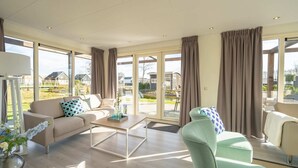 En rummelig stue med en beige sofa, en grøn lænestol og et sofabord. Store vinduer giver udsigt til det udenfor.