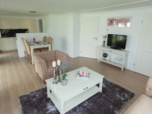 A modern living room with a beige sofa, a white coffee table with candles, a TV on a white stand, and a dining area in the background. - Modern Chalet With Garden, Terrace (Brunssum)