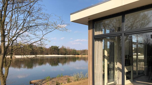 A modern building with large glass windows overlooking a calm lake and a wooden deck. - Modern Chalet With Garden, in Green Kempen (Mol)