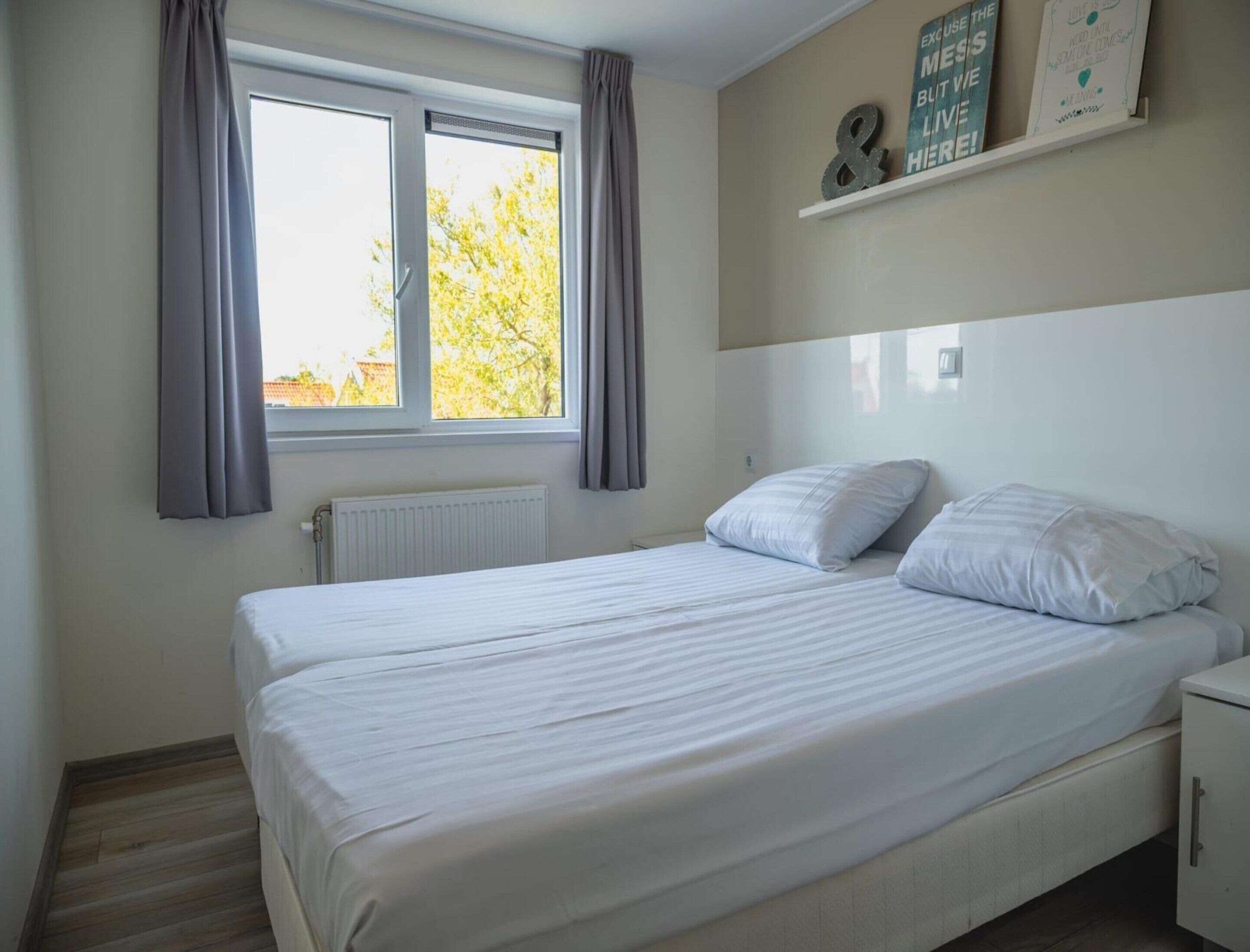 Une chambre à coucher avec un lit, une fenêtre avec des rideaux, un radiateur, une étagère ornée de citations inspirantes et une armoire blanche.