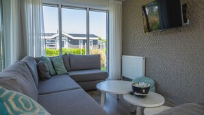 A modern living room with a sectional sofa, a round coffee table, a flat-screen TV mounted on the wall, and large windows offering a view of a residential area. - Modern, Spacious, Detached Holiday Home, Near Alkmaar (West-Graftdijk)