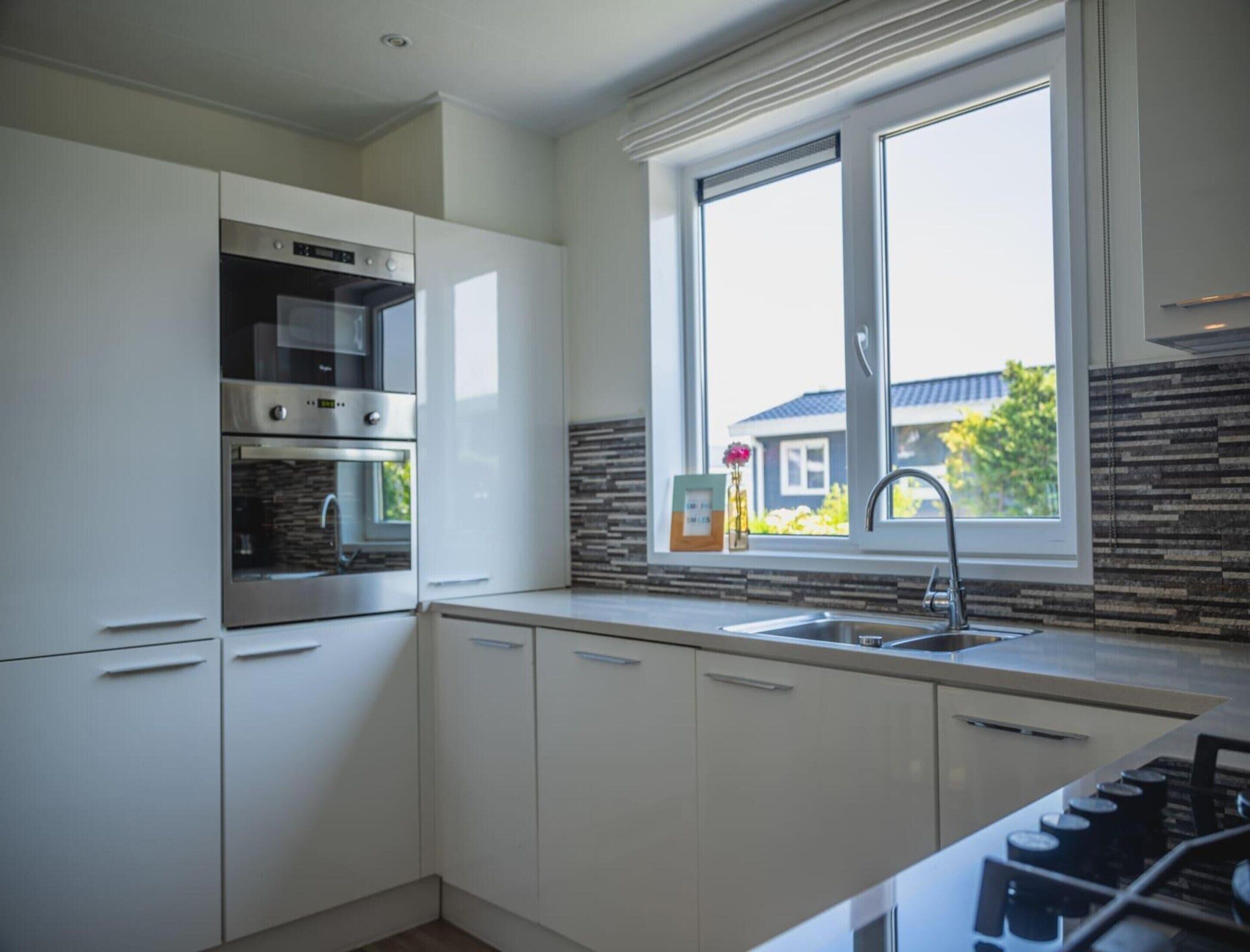 Eine moderne Küche mit weißen Schränken, einem Einbauofen, einer Mikrowelle und einem großen Fenster mit Blick auf ein Haus.