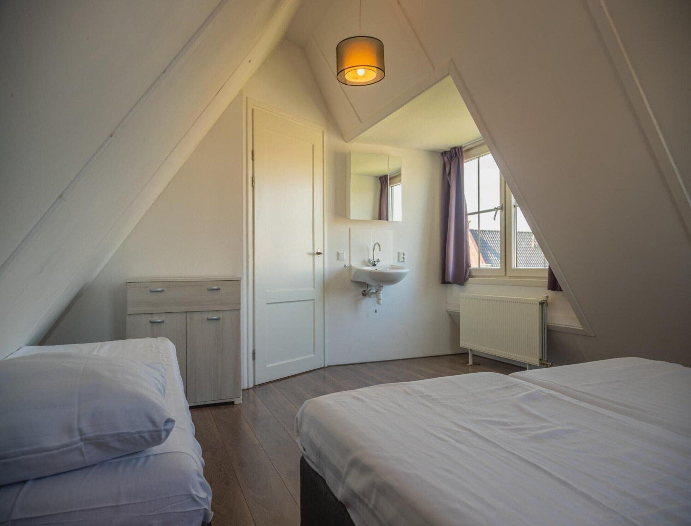 A bedroom with a sloped ceiling, a bed, a dresser, a sink, and a window with curtains.