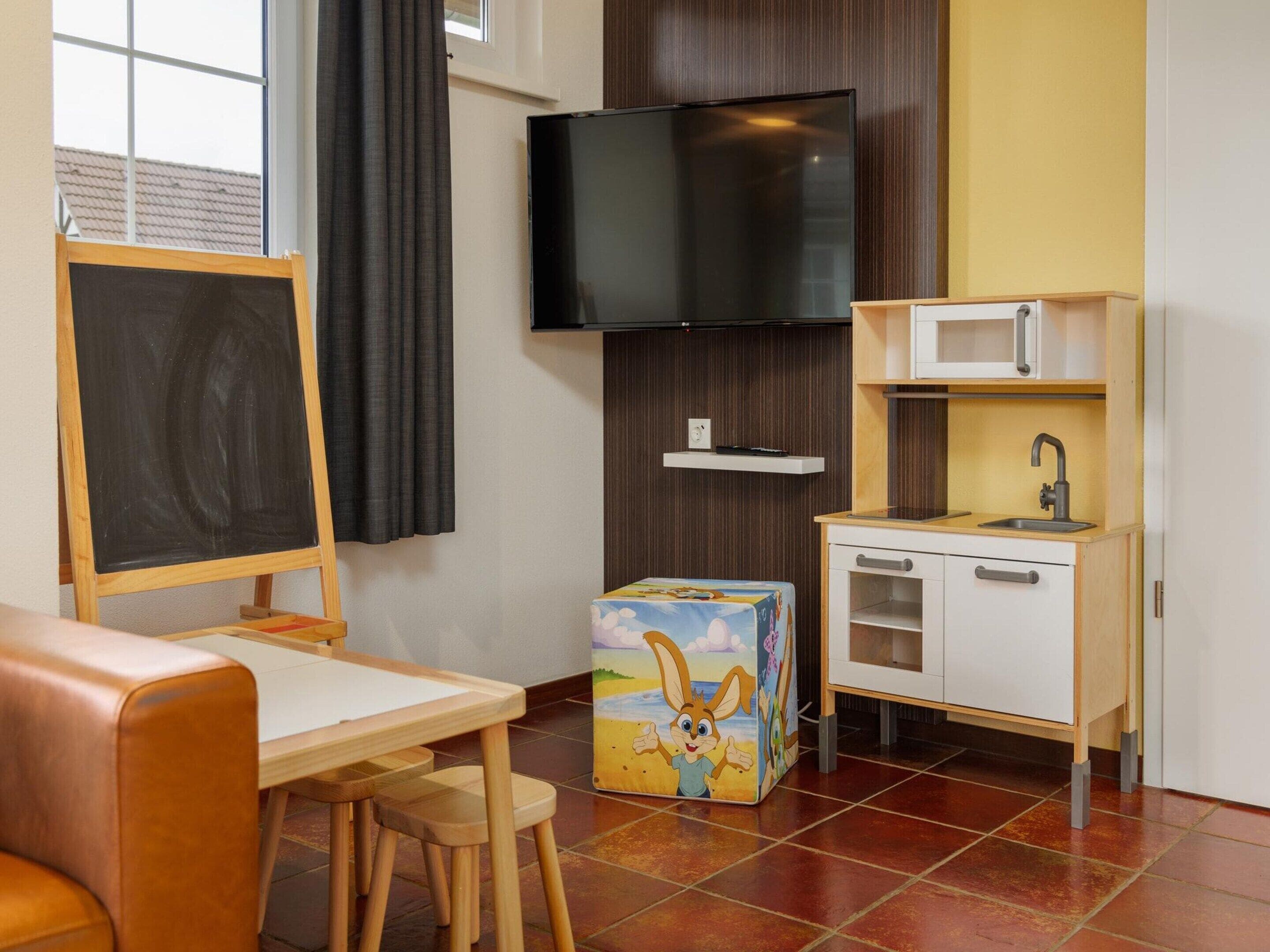 Une pièce comprenant un bureau en bois, un tableau noir, une télévision, un four à micro-ondes et une petite kitchenette.