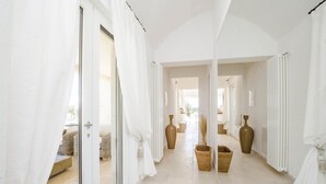 Un pasillo luminoso con paredes y puertas blancas, que conduce a una zona de estar con mobiliario de mimbre.