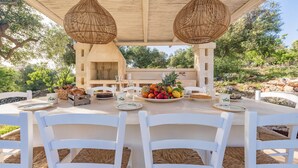 Un área de comedor al aire libre con una mesa dispuesta para cuatro personas, rodeada de sillas blancas, bajo una pérgola de madera con lámparas de mimbre colgantes.