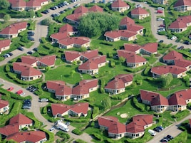 An aerial view of a residential area with multiple houses, each with a red roof, surrounded by well-maintained lawns and parked cars.