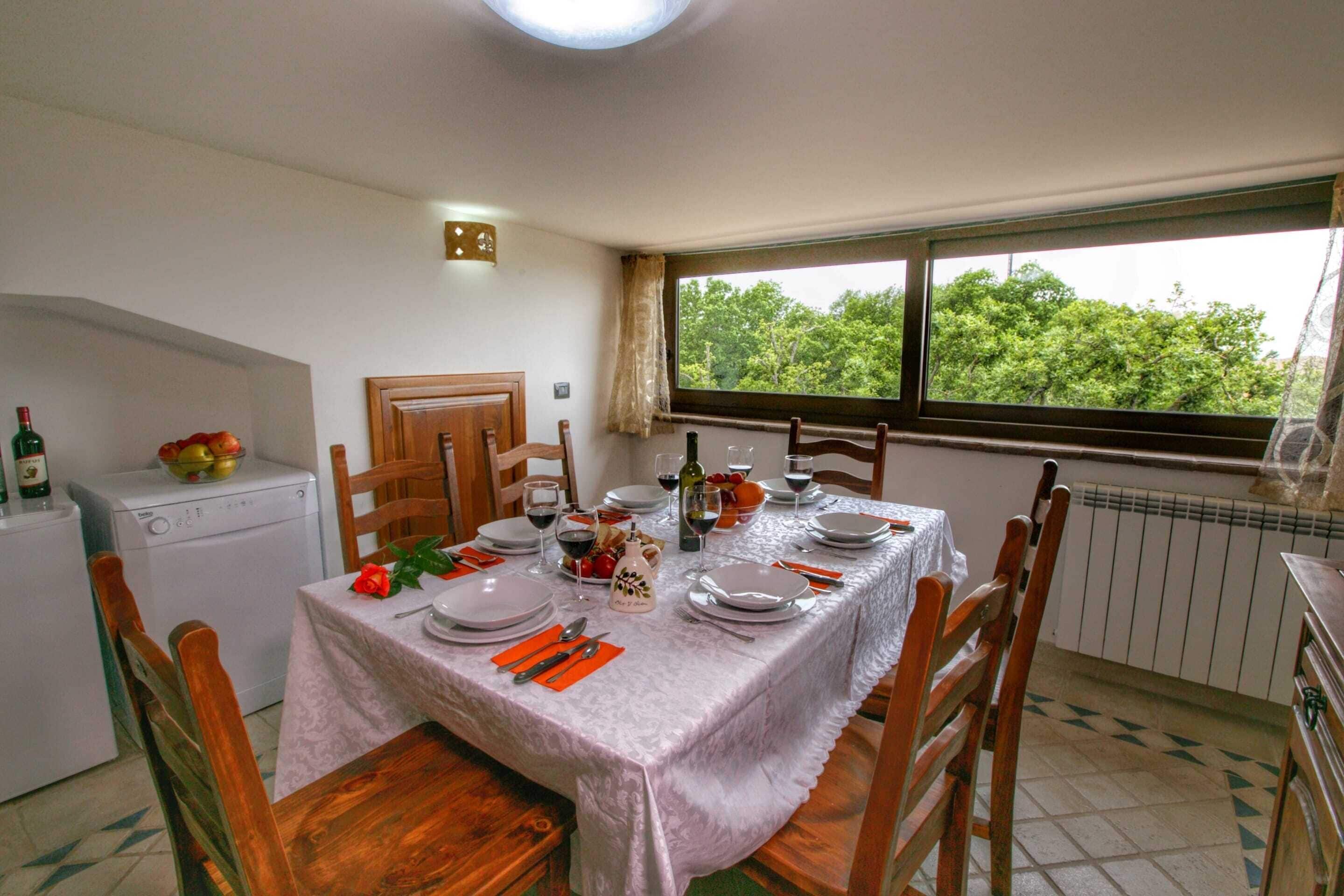 A dining area with a table set for four, chairs, a window with a view of trees, a radiator, and a small kitchen area with a washing machine.