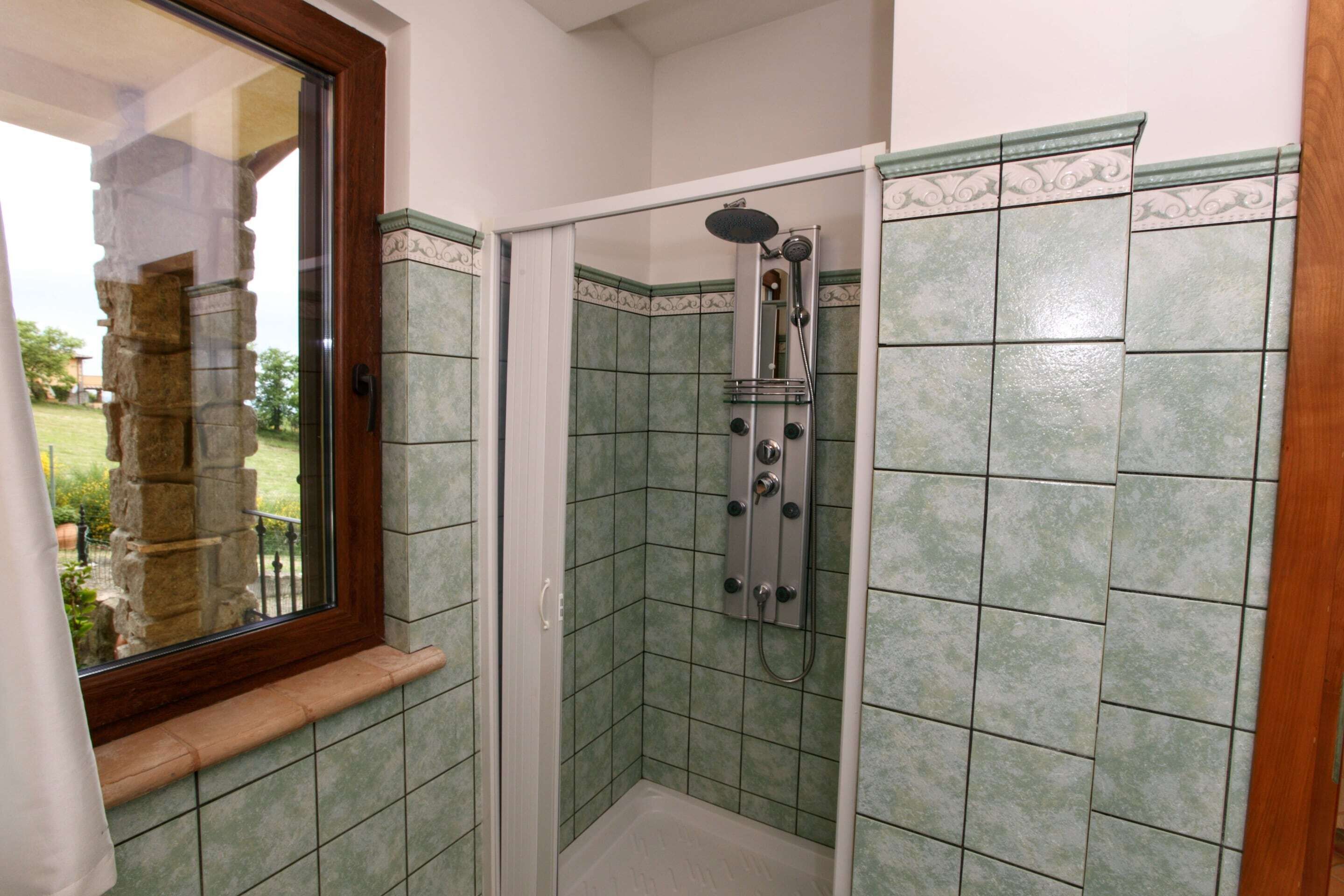 A shower with a glass door, green tiles, and a window with a wooden frame.
