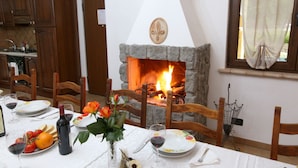 Un coin repas avec une cheminée en pierre, des chaises en bois, une table dressée pour le repas et, au fond, une cuisine.