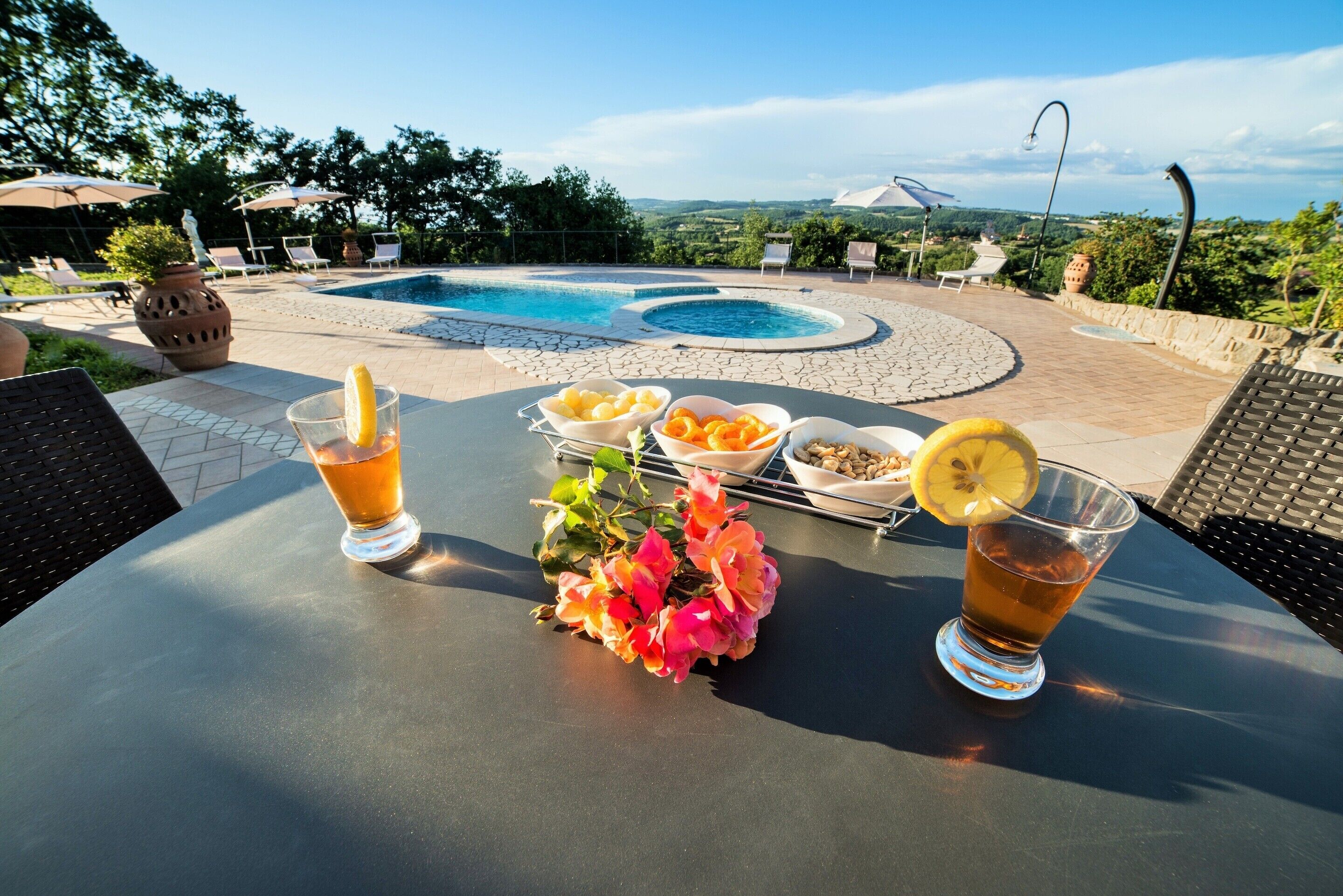 A table with drinks and fruit bowls, a pool, and outdoor seating with umbrellas.