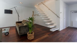 A modern living room with a dark wood floor, a grey sofa, a floor lamp, a potted plant, and a staircase with white steps. - Beautiful Well Equipped Apartment Near the Beach (Domburg)