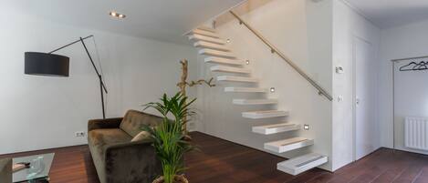A modern living room with a dark wood floor, a grey sofa, a floor lamp, a potted plant, and a staircase with white steps.