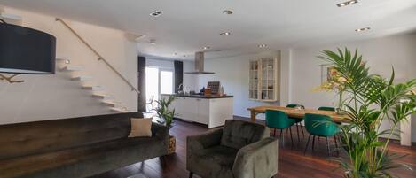 En moderne stue med sofa, lænestol og sofabord, et spiseområde med stole, et køkken i baggrunden og en trappe.