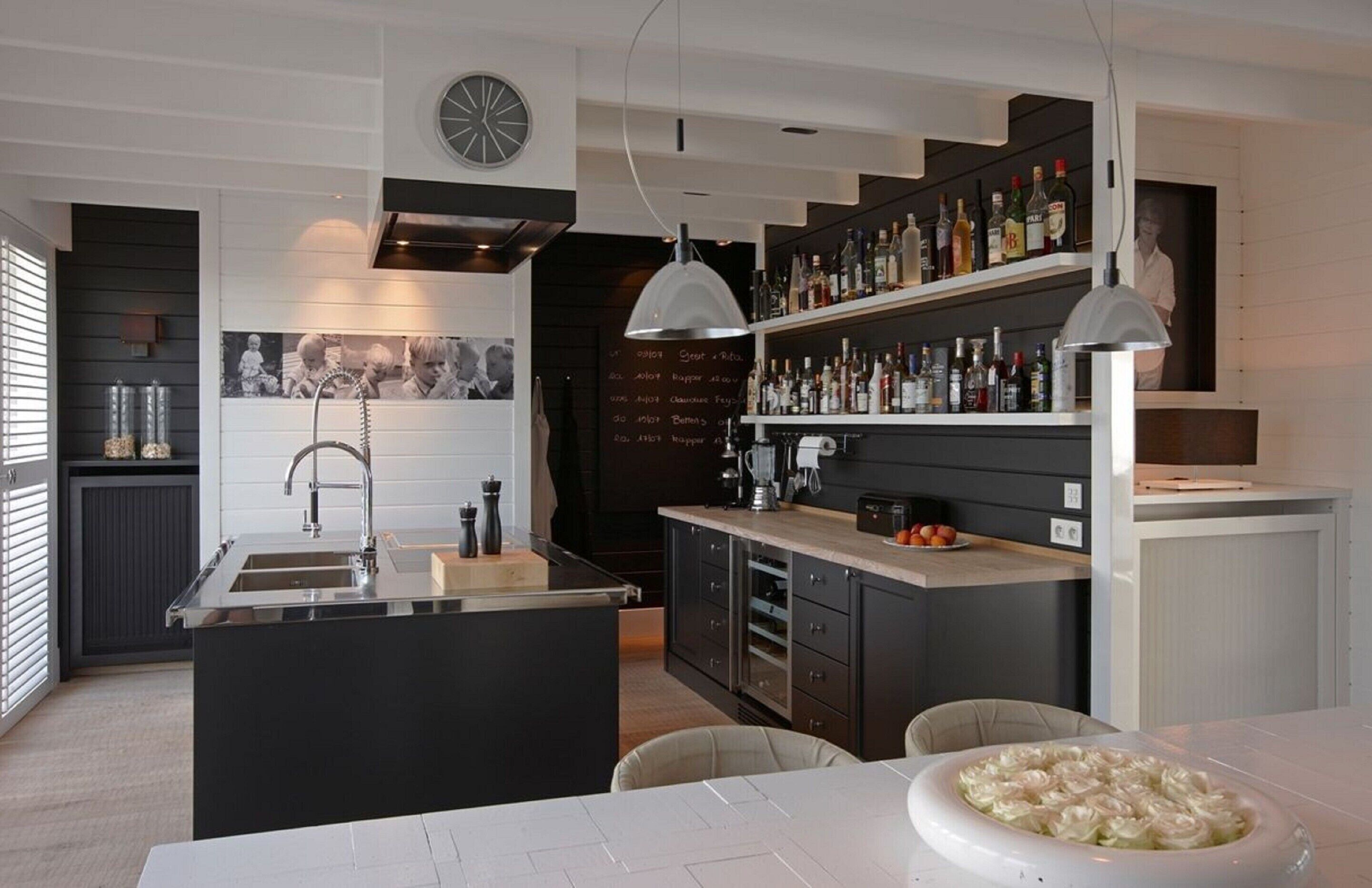 Une cuisine moderne dotée d’un îlot central, d’un coin bar avec des étagères remplies de bouteilles et d’un espace repas avec une table et des chaises blanches.