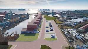 Vista aerea di una marina con barche ormeggiate, un parcheggio con auto e una distesa d'acqua sullo sfondo.