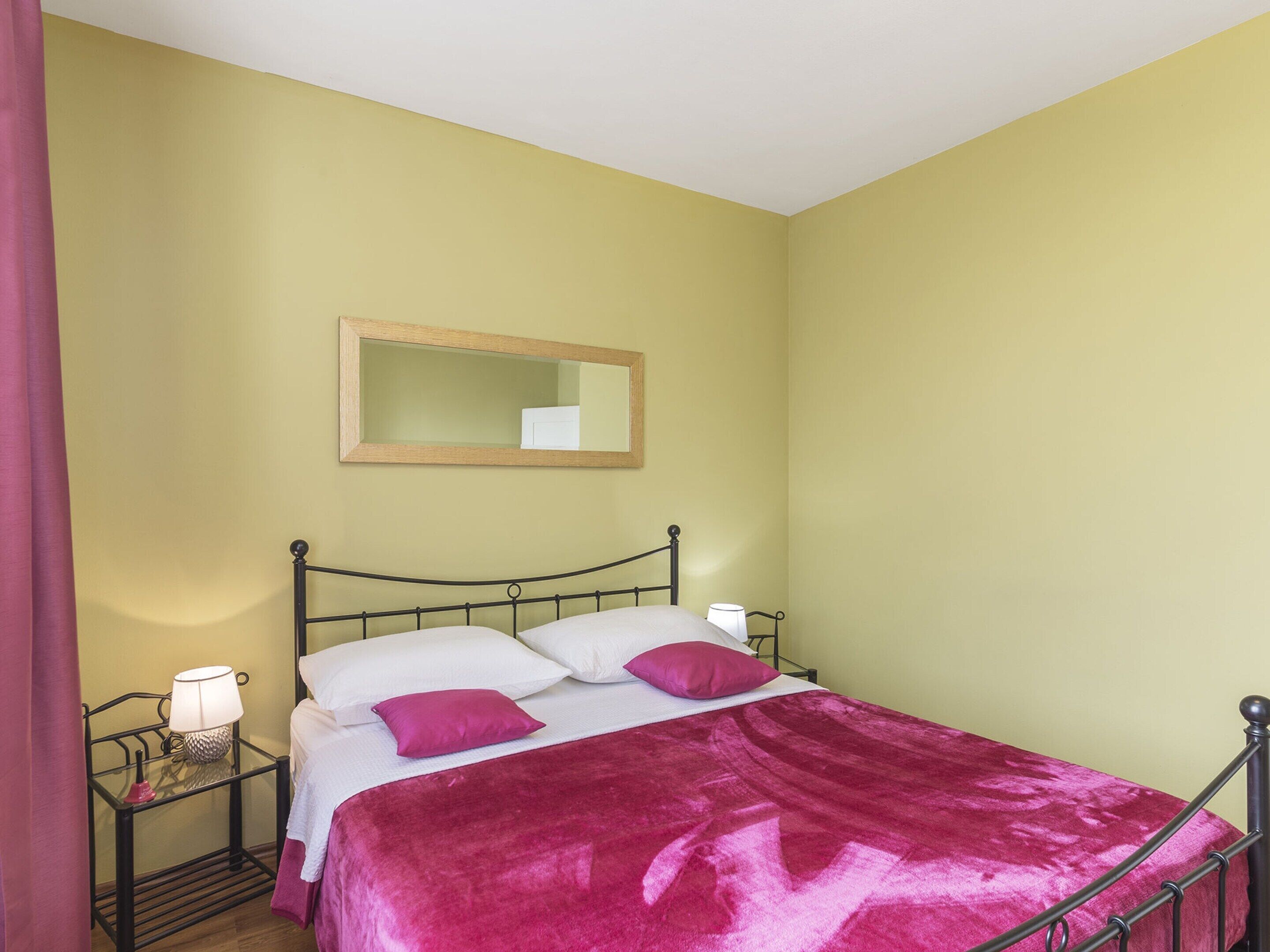 A single bed with a metal frame, a wooden headboard, and a pink bedspread. There is a bedside table with a lamp and a mirror on the wall above the bed.