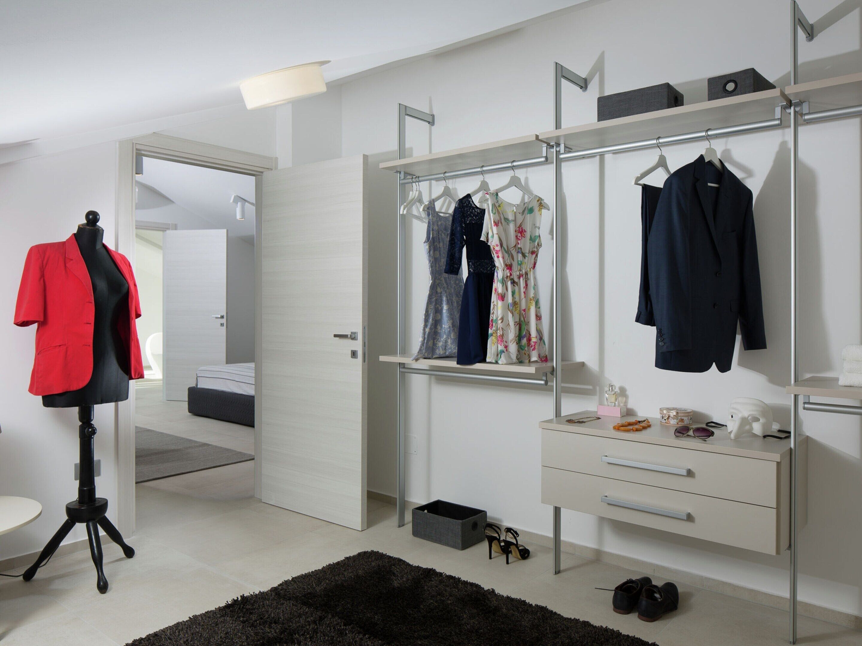 A modern bedroom with a large wardrobe, a vanity with drawers, a mirror, and a mannequin dressed in a red blazer.
