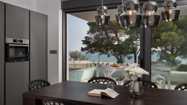 Una cocina moderna con horno integrado, una mesa de comedor con un libro y un jarrón de flores, y vista al mar a través de amplias ventanas.