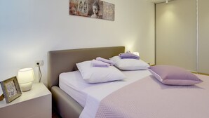 Un lit bien fait, avec une literie blanche et lavande, une lampe de chevet et un cadre photo posé sur une table de chevet.