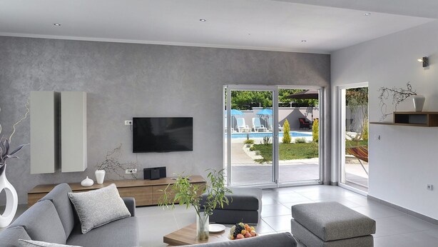 Ein modernes Wohnzimmer mit einem grauen Sofa, einem Couchtisch mit Obst, einem Flachbildfernseher und einer Glastür, die zu einem Poolbereich führt.