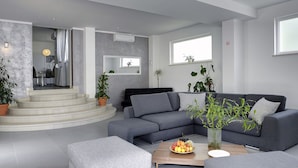 Ein modernes Wohnzimmer mit einem grauen Sofa, einem Hocker und einem Couchtisch. Üchtige Pflanzen und eine Treppe, die in einen Essbereich führt, sind ebenfalls zu sehen.