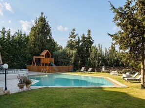 A swimming pool with a wooden play structure, lounge chairs, and a fenced area with trees. - Luxury Villa Near Sea and City, Ping Pong, Childrens Area and BBQ (Rethymno)