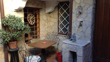 A rustic outdoor seating area with a stone wall, a wooden table, metal chairs, a potted plant, and a small sink.