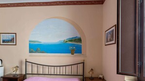 Une chambre à coucher comprenant un lit, une table de chevet, une lampe et une fenêtre ornée d’un tableau représentant un paysage côtier.