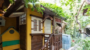 Une maison rustique en bois, à l’extérieur jaune, avec un escalier en bois et une petite construction dotée d’un toit végétalisé, qui ressemble à un nichoir.