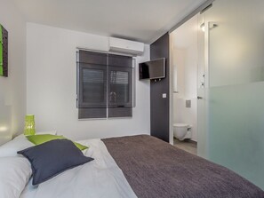 Un dormitorio moderno con cama, televisor montado en la pared y baño con inodoro y lavamanos.
